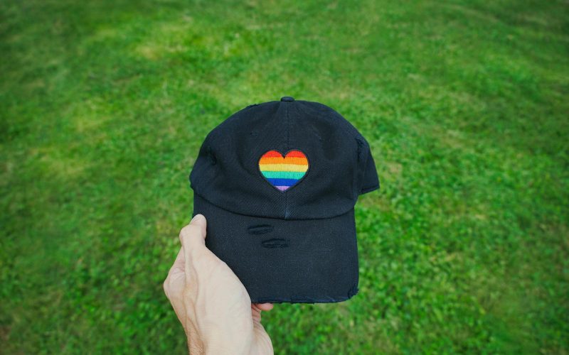 Hand holding a black cap with a rainbow heart, symbolic of pride, in a grassy field.