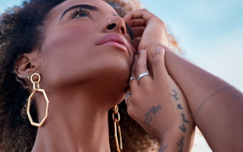 Portrait of a woman with tattoos and gold jewelry gazing thoughtfully into the sky.