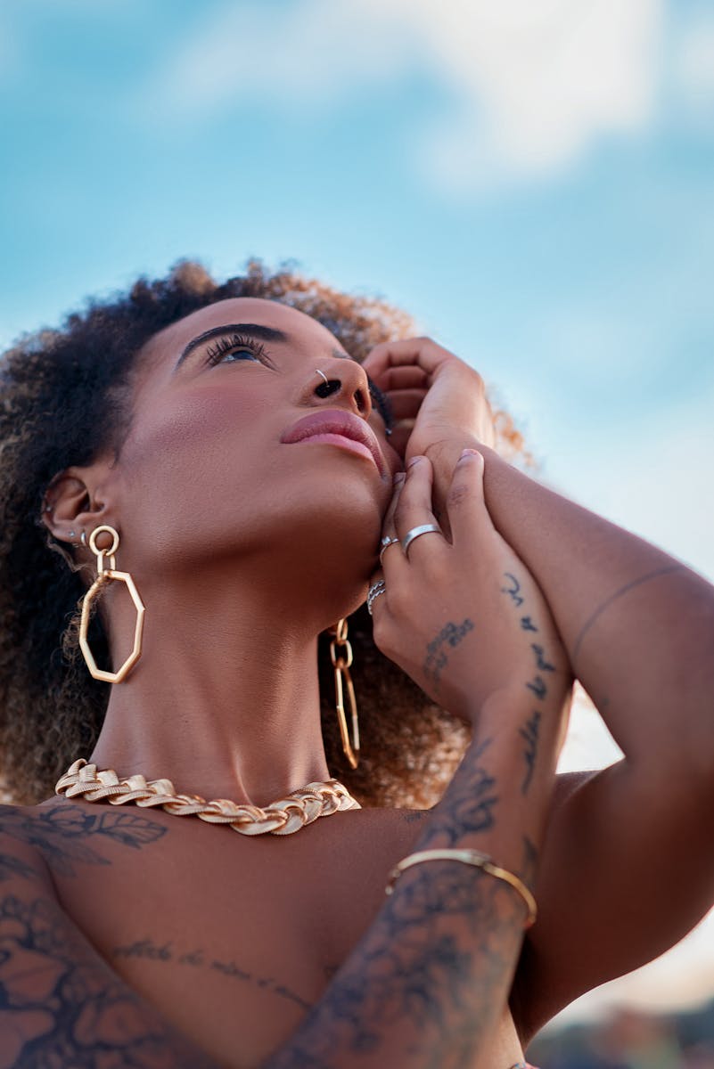 Portrait of a woman with tattoos and gold jewelry gazing thoughtfully into the sky.